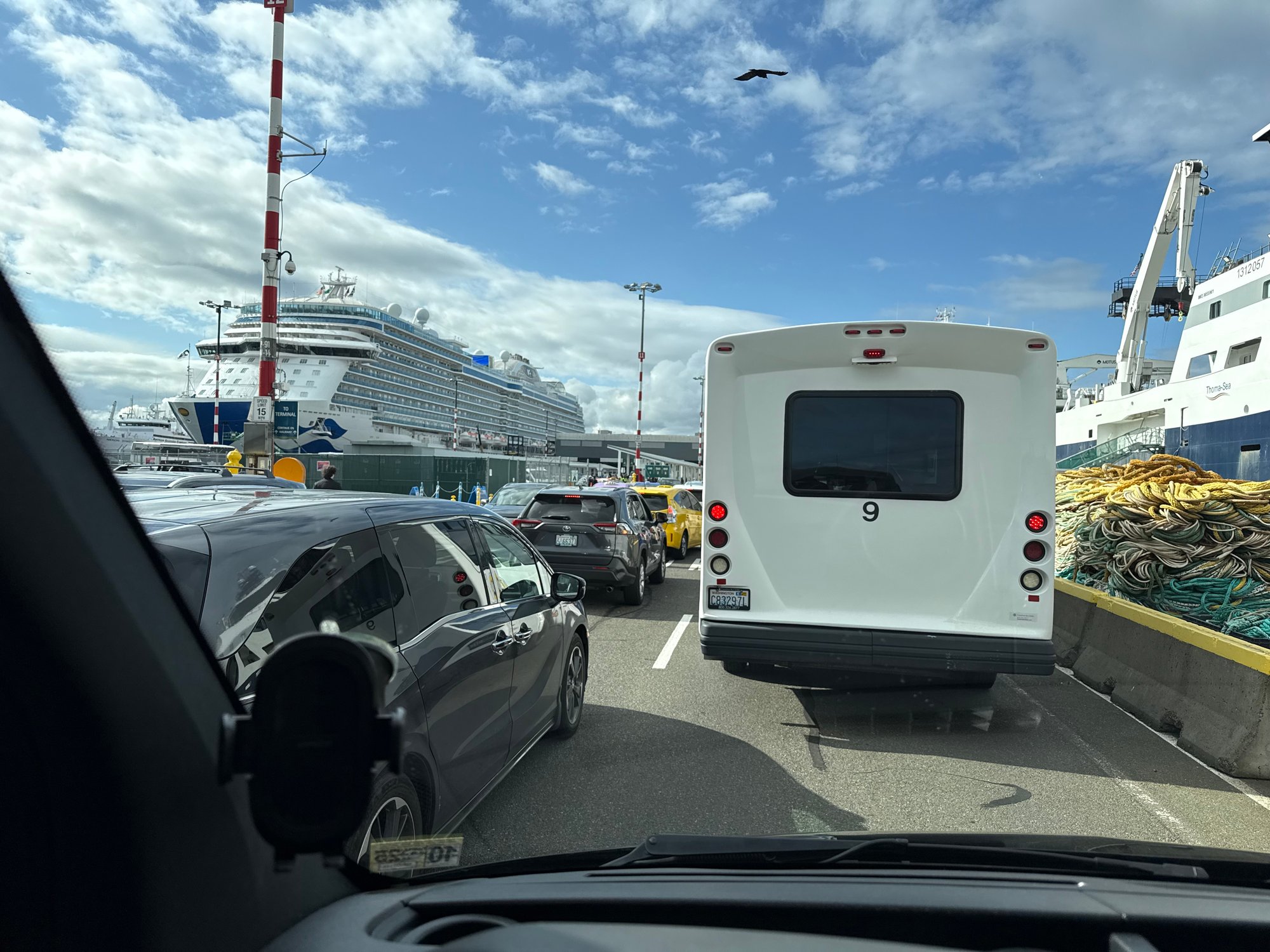 View from a car in port traffic, showing a cruise ship, cargo ship, bus, and other vehicles.