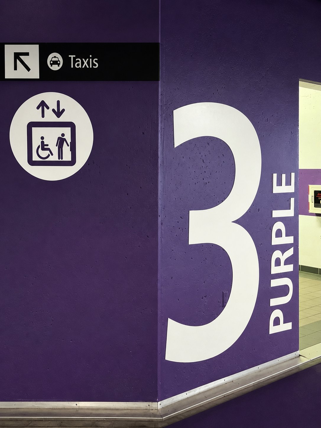 A vibrant purple wall displaying a large number 3, with signs for taxis and an accessible elevator.