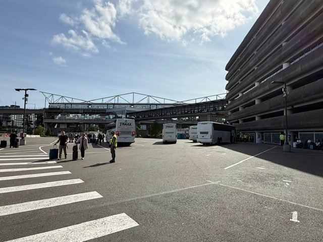 Travelers with luggage near white shuttle buses and a multi-story building under a blue sky.