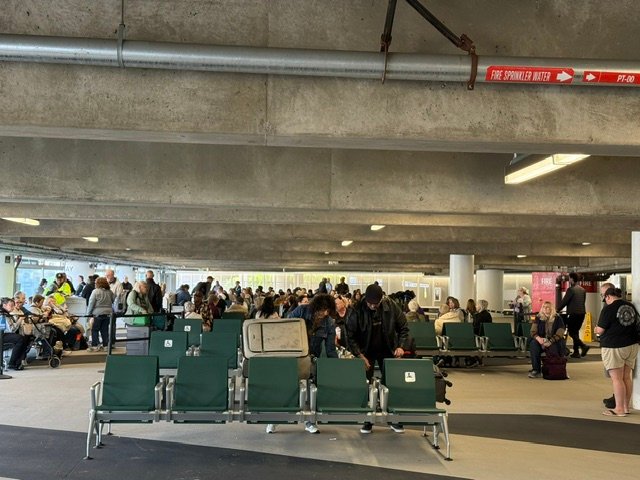 A bustling indoor waiting area with many travelers sitting on green chairs and standing with luggage.